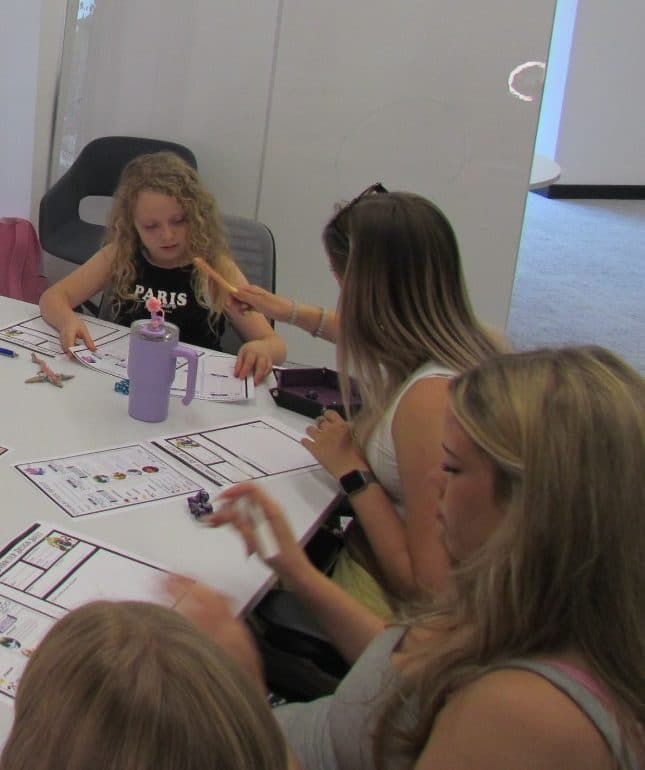 A girl and two young women sitting around a table with character sheets and dice during a Role Resolve storytelling session.