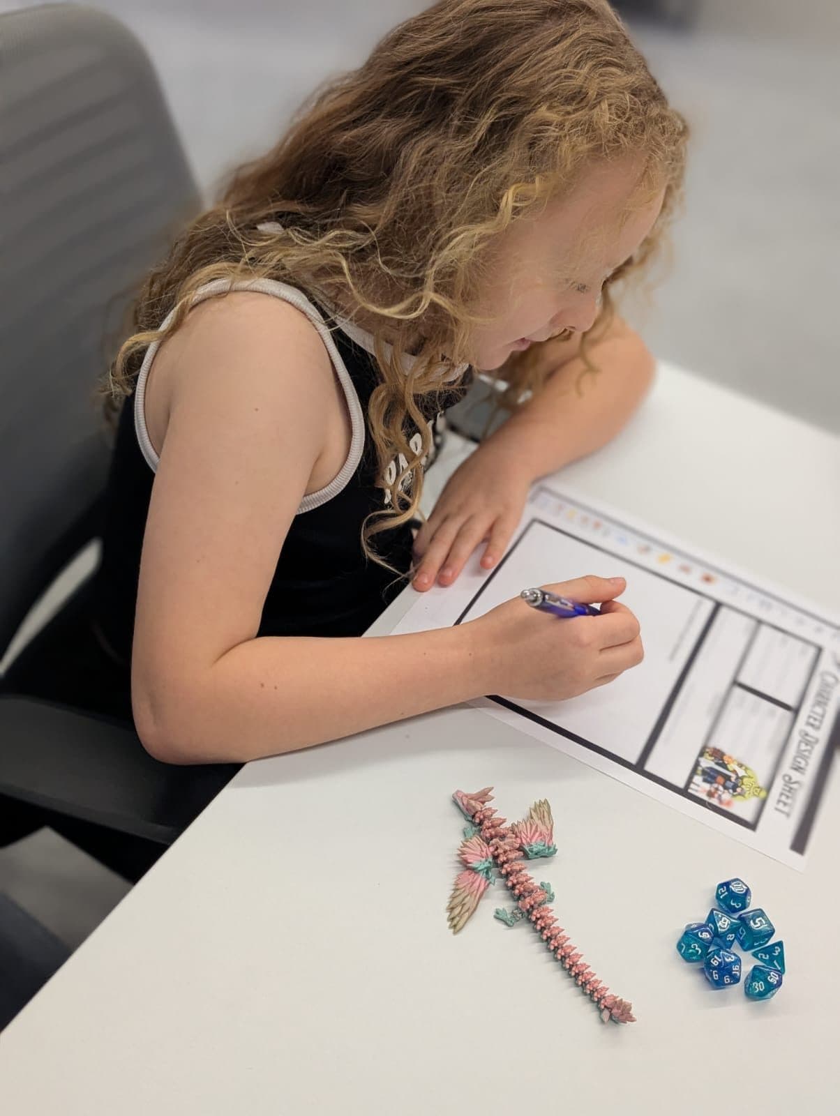 A girl drawing on a character sheet, with dice and a toy dragon on the table beside her